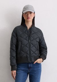 Czarna pikowana kurtka bomber z zakładanym na przód zamkiem, ściągaczami przy mankietach i kołnierzu; noszona z szarą haftowaną czapką i niebieskimi dżinsami.