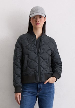 Czarna pikowana kurtka bomber z zakładanym na przód zamkiem, ściągaczami przy mankietach i kołnierzu; noszona z szarą haftowaną czapką i niebieskimi dżinsami.