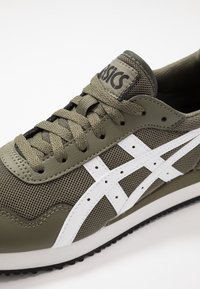 Buty sportowe w oliwkowo zielonym kolorze z białymi akcentami. Wykonane z siateczki i skóry, z zaokrąglonym noskiem, płaskimi sznurowadłami i gumową podeszwą.