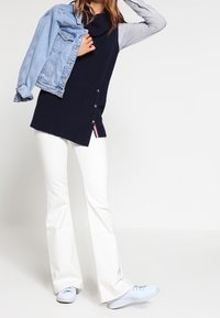 Námořnický pletený svetr s rolákem a knoflíkovými detaily, v kombinaci s bílými rozšířenými džíny a světle modrou denimovou bundou, nošený s bílými teniskami.