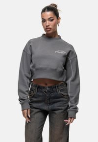 Felpa grigia accorciata con colletto alto e logo ricamato bianco, abbinata a jeans in denim scuro. Vestibilità ampia e texture morbida.