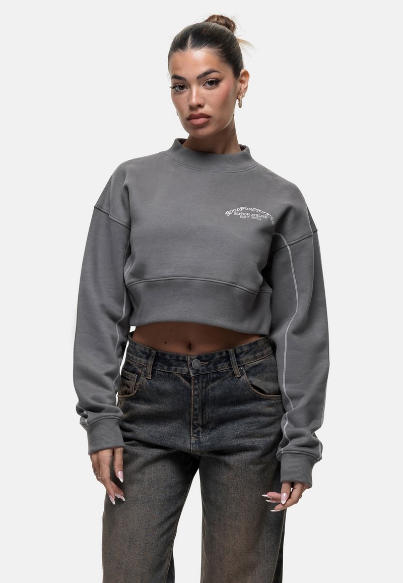 Felpa grigia accorciata con colletto alto e logo ricamato bianco, abbinata a jeans in denim scuro. Vestibilità ampia e texture morbida.