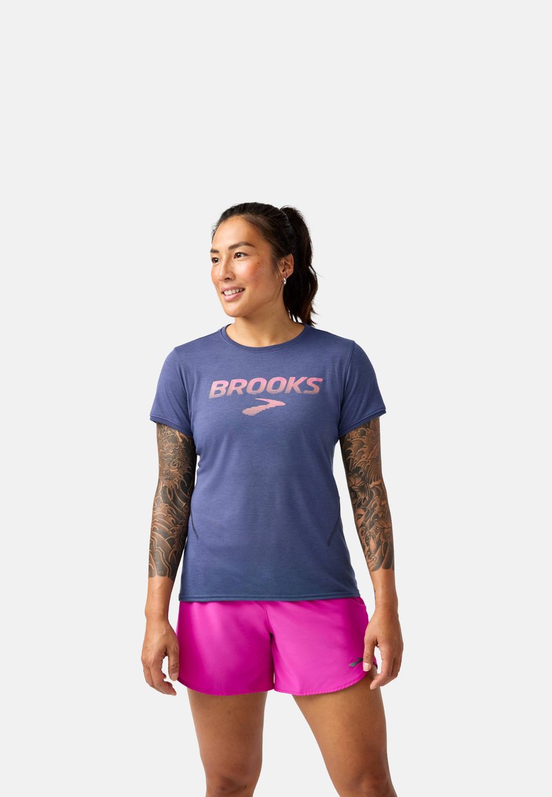 Camiseta deportiva azul marino de manga corta con el logotipo rosa "BROOKS", combinada con pantalones cortos de un brillante color rosa. Tejido suave, corte moderno, estilo casual.