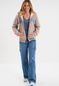 Felpa con cappuccio beige foderata in pile, con grafiche blu. Abbinata a pantaloni cargo in denim blu a vita alta e scarpe da ginnastica bianche.