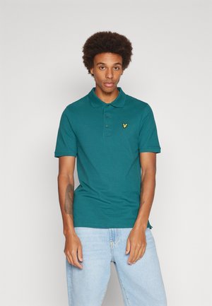 Polo shirt color teal con patta a due bottoni e un piccolo logo giallo. Indossato con jeans azzurri, con maniche corte e vestibilità regolare.