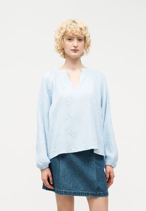 Blondínka v svetlomodrej blúzke s dlhým rukávom a očkovými detailmi a tmavomodrej denimovej sukni na jednoduchom pozadí.