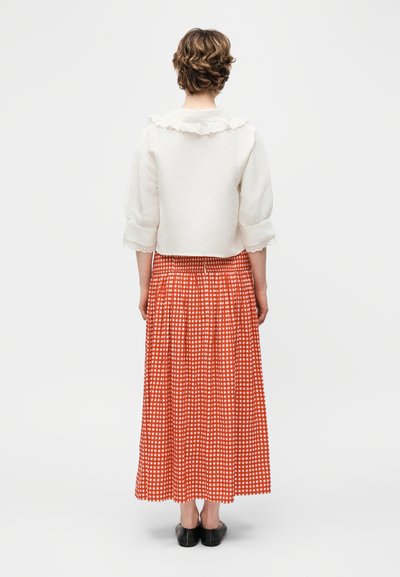 Άτομο στέκεται με την πλάτη γυρισμένη, φοράει λευκό πουκάμισο με βολάν στον γιακά και τις μανσέτες, μακριά κόκκινη και λευκή καρό φούστα και μαύρα παπούτσια.