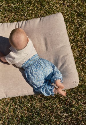 Bébé couché sur le ventre sur un coussin beige à l'extérieur sur l'herbe, portant une chemise blanche et un pantalon bleu à fleurs.