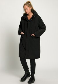Femme portant un long manteau rembourré noir avec une capuche, un jean noir et des bottes noires imposantes, se tenant avec les mains dans les poches du manteau.