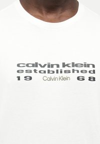 Κοντινή λήψη ενός ατόμου που φοράει λευκό μπλουζάκι με το κείμενο «calvin klein established 1968» τυπωμένο στο στήθος με μαύρα και ανοιχτό πράσινα γράμματα.