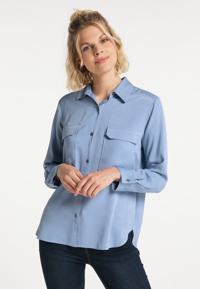 Young woman with wavy blonde hair wearing a light blue button-up shirt with pockets and dark jeans, standing against a white background.