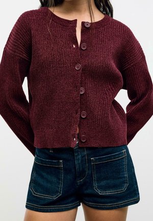 Femme portant un cardigan côtelé bordeaux boutonné et un short en jean bleu foncé avec des poches avant, visible du cou jusqu'à mi-cuisse.