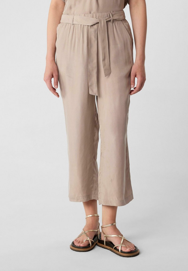 Beige, knöchellange Hosen mit einem Bindegürtel und weitem Schnitt. Leichter Stoff mit glatter Textur, kombiniert mit Riemchensandalen.