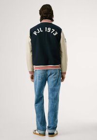 Mornarsko modra in smetana jakna z rdečimi trakovi ob ovratniku, z vezenim napisom "PJL 1973" na hrbtu. Ujemajoči se z svetlo modrimi jeans hlačami.
