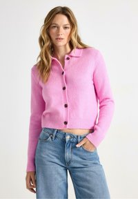 Rosa ullkofta med krage, med bruna knappar, ribbat avslut och långa ärmar. Matchat med högmidjade blå jeans.