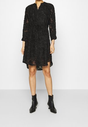 Czarna sukienka z długimi rękawami, dekoltem w serek i paskiem w talii. Przezroczysta tkanina z fakturą kwiatowego wzoru, długość do połowy uda, zestawiona z czarnymi botkami za kostkę.