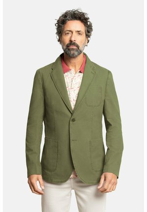 Blazer verde oliva hecho de una tela de textura suave. Presenta dos bolsillos frontales y un cierre de un solo botón, llevado sobre una camisa estampada.