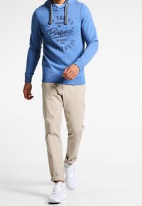 Blå hoodie med grå dragkedjor och tryckt text, tillsammans med ljusbeiga byxor och vita sneakers, vilket skapar en avslappnad outfit.