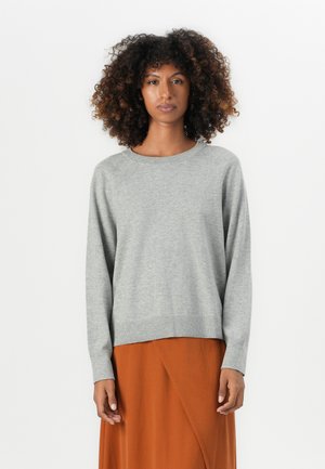 Vrouw met krullend haar, gekleed in een lichtgrijze lange mouwen trui en een roestoranje rok, staand voor een effen witte achtergrond.