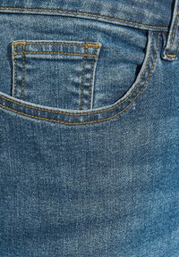 Niebieskie dżinsy z widoczną haftowaną kieszenią z tyłu w kolorze pomarańczowym. Tkanina ma wyblakłe, teksturowane wykończenie.