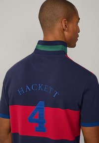 Polo de color marino con rayas horizontales rojas y azules, con el número "4", el bordado "HACKETT" y un acento en el cuello verde.