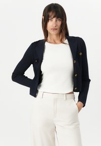 Námořnický vroubkovaný blazer se zlatými knoflíky a klopami, nošený přes bílý vroubkovaný top. V kombinaci se světlými tailored kalhotami.