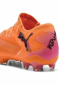 Scarpino da calcio arancione brillante con tomaia testurizzata, logo Puma nero e accenti a gradiente rosa sul tallone. Tacchetti trasparenti sulla suola per trazione.