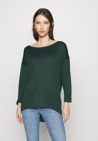 Mørkegrøn, langærmet top med en bred halsudskæring, afslappet pasform og asymmetrisk kant. Blødt stof med en glat tekstur. Matchet med lyseblå jeans.