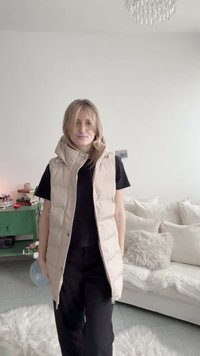 Žena v béžovej prešívanej bezrukávovej bunde cez čierne tričko a nohavice stojí v jasnej obývačke so zelenou skrinkou a bielou sedačkou.