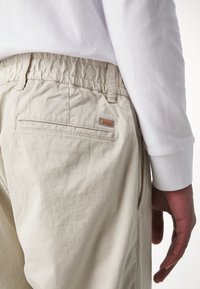 Chinos beige claro con cinturilla elástica, bolsillo lateral y un bolsillo trasero. Presenta un pequeño logo marrón de "BOSS" en la parte posterior. Camisa blanca de manga larga visible.