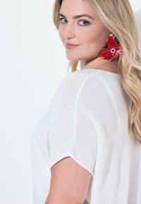 Top bianco, leggero, con maniche corte e vestibilità rilassata. Presenta grandi orecchini a forma di fiore rossi, che aggiungono un contrasto di colore deciso.