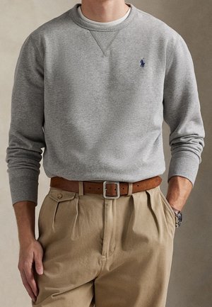 Mand iført en grå sweatshirt med et lille logo, beige plisserede bukser, brun læderbælte og sølv armbåndsur med den ene hånd i lommen.