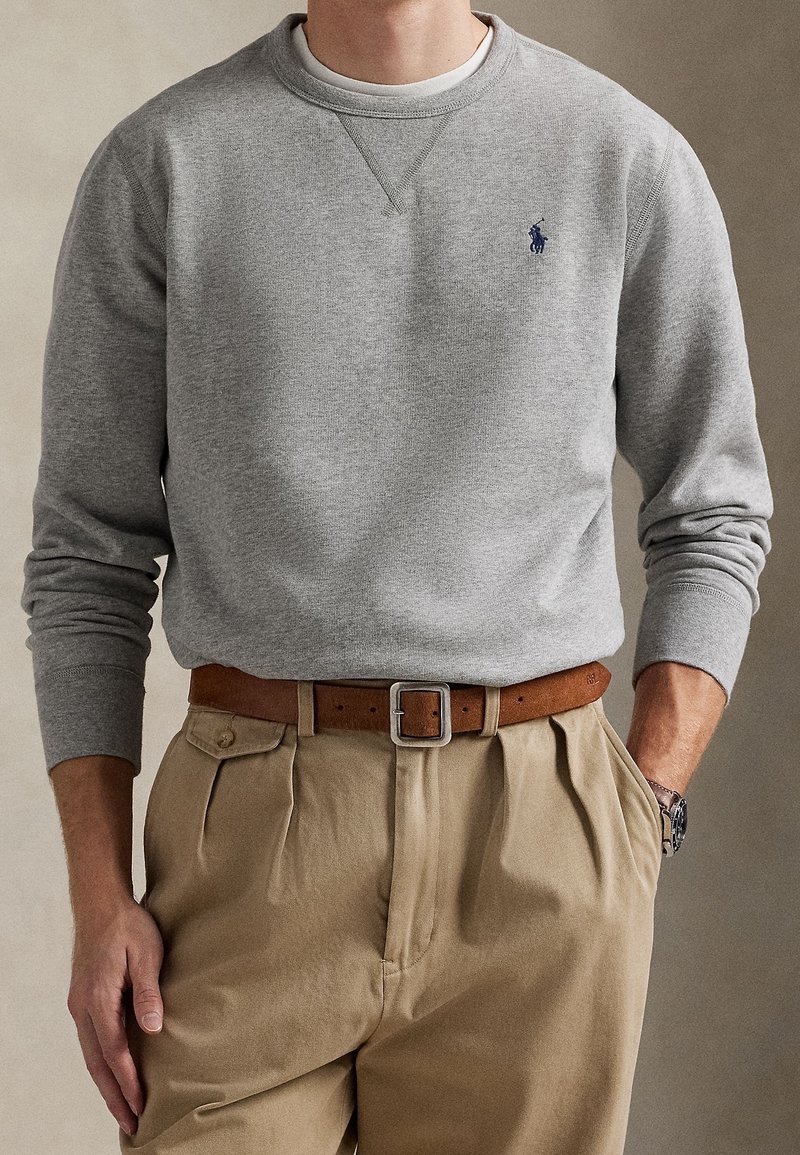 Homme portant un sweat-shirt gris avec un petit logo, un pantalon plissé beige, une ceinture en cuir marron et une montre-bracelet argentée, une main dans la poche.