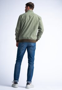 Olivově zelená bomber bunda s hnědým rib kálem, nošená přes modré džíny. Obsahuje stojáček a viditelné švy.