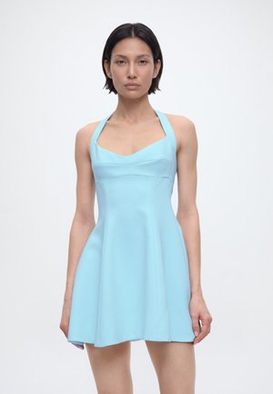 Γυναίκα με κοντά σκουρόχρωμα μαλλιά, ντυμένη με αμάνικο φωτεινό γαλάζιο φόρεμα με halter λαιμόκοψη, σε απλό λευκό φόντο.