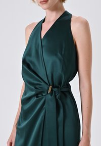 Dunkelgrünes Satinabendkleid mit einem Halterdesign, das eine verdrehte Taille und eine goldfarbene Akzentuierung an der Seite aufweist. Glatte, fließende Textur.