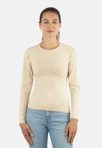 Frau mit dunklem, nach hinten gebundenem Haar, die goldene Ohrringe, ein beiges langärmeliges, figurbetontes Shirt und hellblaue Jeans trägt, vor einem schlichten Hintergrund.