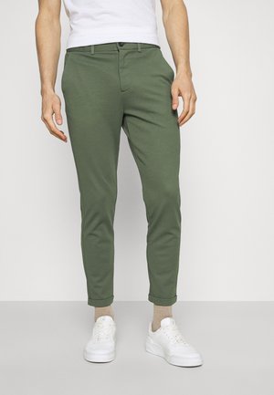 Hombre que lleva pantalones ajustados color verde oliva remangados en los puños, calcetines beige, zapatillas blancas y una camiseta blanca, de pie frente a un fondo liso.