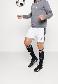 Personne portant un sweat à capuche gris, un short Adidas blanc, des chaussettes noires rayées et des crampons noirs, contrôlant un ballon de football en plein air sur un fond uni.