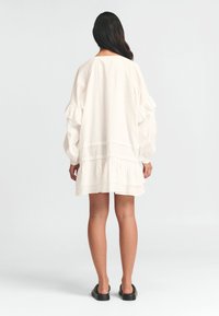 Valge kihiline kleit ruffli varrukatega ja tekstuurse kangaga. Omab vabalt langevat lõiget ja kõrget kaelust, ning on kombineeritud mustade lameede sandaalidega.