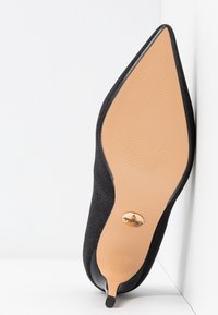 Chaussure à talons hauts en tissu noir avec un bout pointu, surface texturée et une semelle beige lisse ornée d'un accent logo doré.