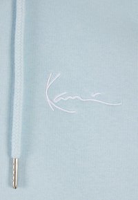 Lys blå genserstoff med en glatt tekstur, med en hvit brodert logo. Inkluderer en lys blå snor med en sølvmetallspiss.