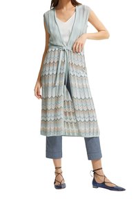 Gilet lungo azzurro chiaro in tessuto semitrasparente a motivo ondulato, con cintura in vita, abbinato a pantaloni in denim corti e ballerine blu.
