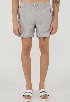 Homme portant un short de bain gris clair avec des coutures latérales rouges et des sandales Emporio Armani blanches, debout devant un fond uni.