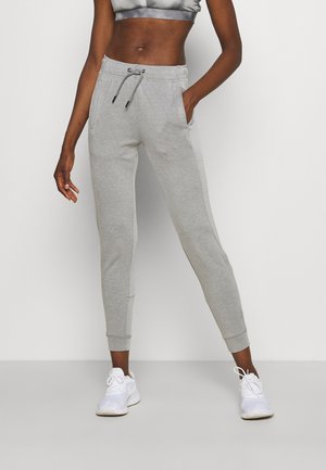 Wąskie, jasnoszare spodnie dresowe typu jogger z wiązanym w pasie ściągaczem, kieszeniami i ściągaczami przy kostkach, noszone z białymi butami sportowymi.
