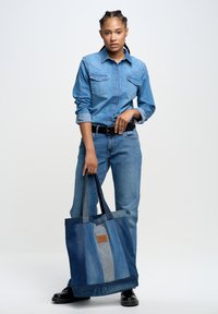 Denim-Shopper mit verschiedenen Blautönen, robustem Stoff, doppelten Tragegriffen und einem Lederakzent. Kombiniert mit einem Denim-Hemd und -Hose.
