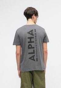T-shirt cinza em algodão com gola redonda, apresentando uma grande impressão preta "ALPHA INDUSTRIES" verticalmente nas costas. Mangas curtas, ajuste descontraído.