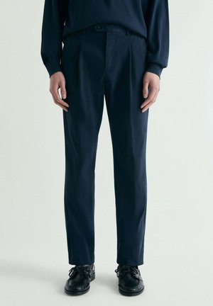 Hombre con pantalones entallados de color azul marino oscuro, zapatos de cuero negros con cordones y un suéter oscuro, sobre un fondo claro y liso.