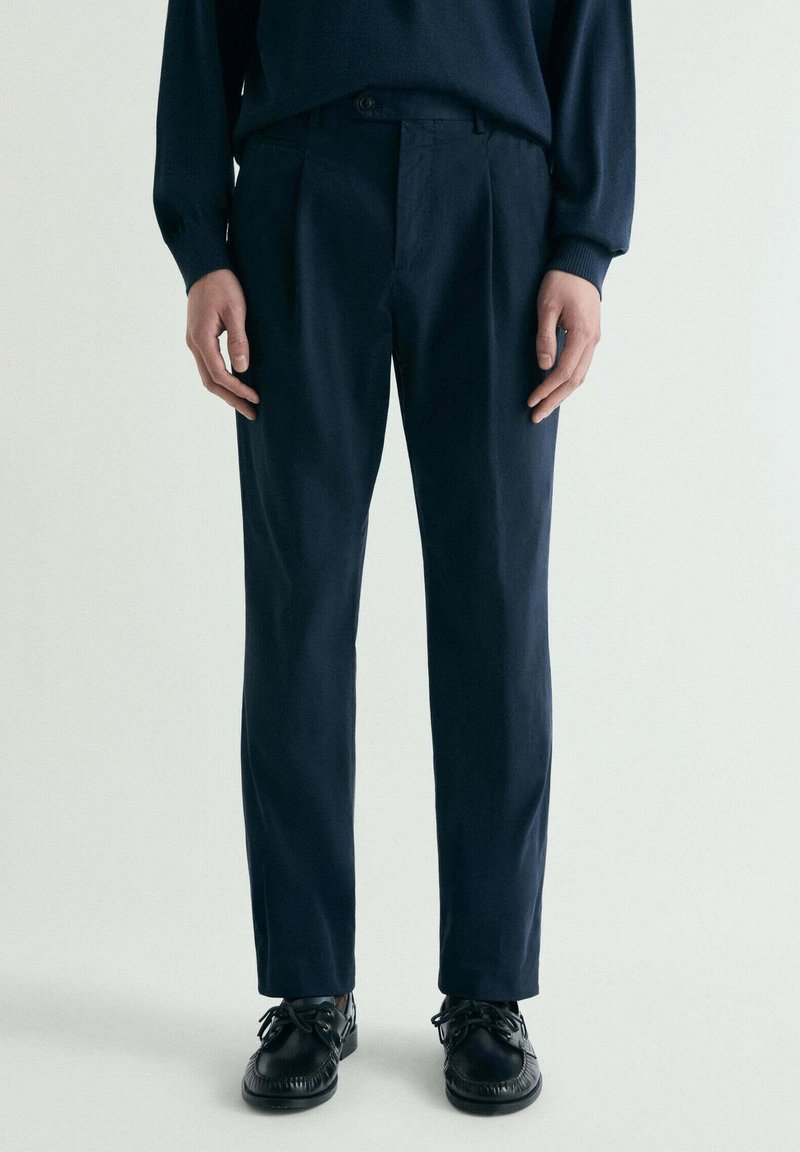 Hombre con pantalones entallados de color azul marino oscuro, zapatos de cuero negros con cordones y un suéter oscuro, sobre un fondo claro y liso.