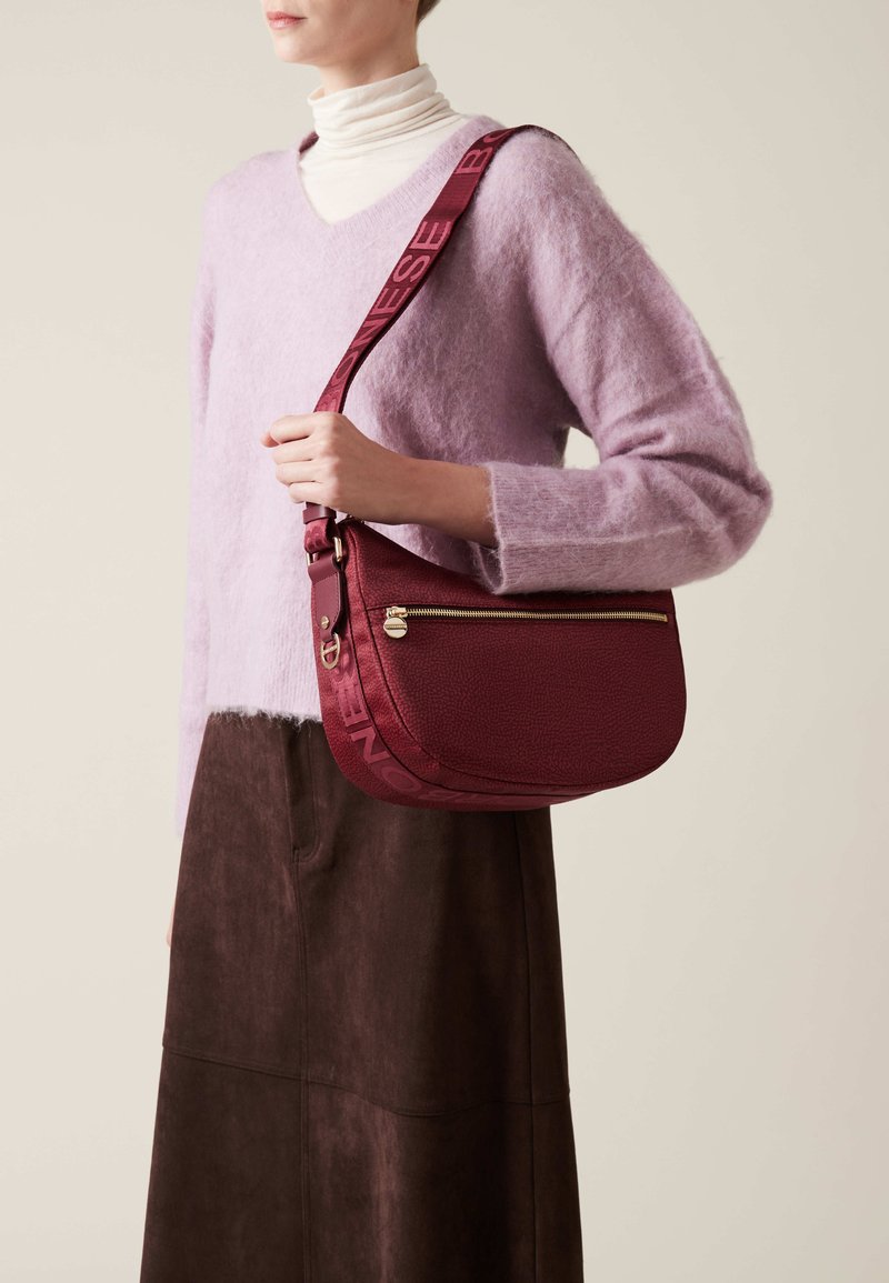 Burgundy leather shoulder bag with textured finish, gold zipper accent, and adjustable strap featuring embossed branding. Worn crossbody.